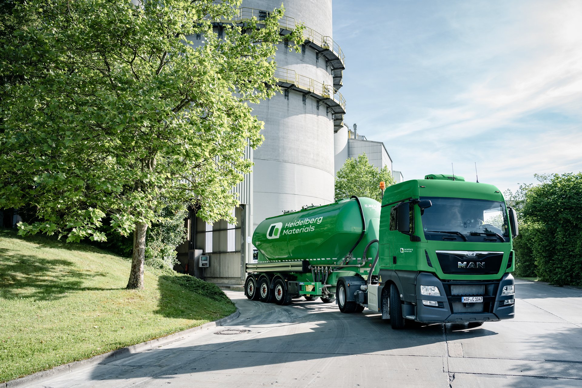 A green silo truck parked in front of a cylindrical building. Trees are visible on the left.